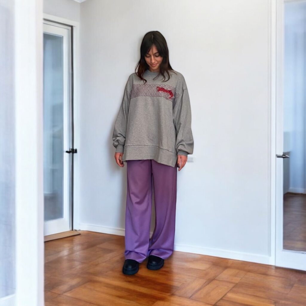 Image of a brunette young woman wearing The Lace Embroidered Sweatshirt by Meagratia brand. The sweatshirt is oversized, in grey colour with a lace band along the fron and a red embroidery. It's paired with a lilac wide leg trousers and comab boots