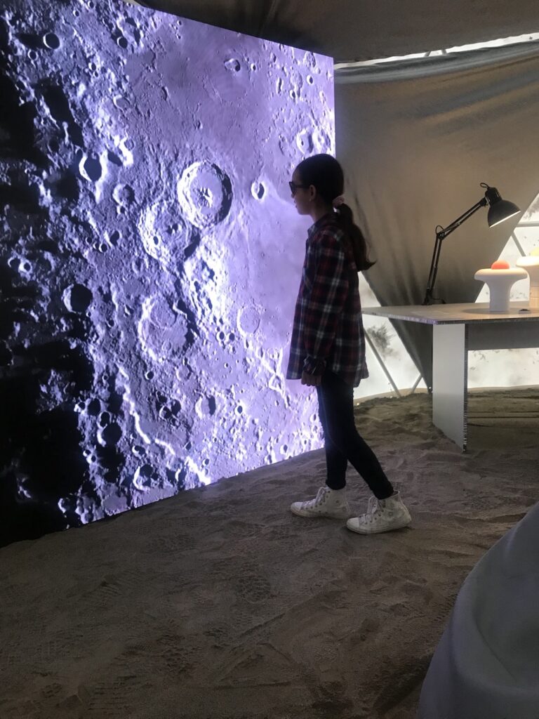 Salone del Mobile: una ragazzina guarda uno shermo su cui scorrono immagini della luna e dei suoi crateri in una installazione che rappresenta il paesaggio lunare.