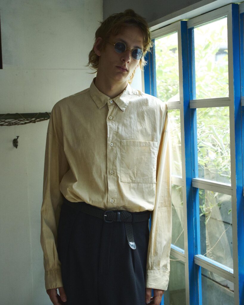 A young man wearing the clay dye beige shirt by GoodNeighbors Shirt. Its earthy, hand-dyed texture visible—paired with tailored black trousers and a slim belt. Round John Lennon-style sunglasses add a retro touch as he leans against a weathered white and blue wooden window, natural light highlighting the shirt’s organic wrinkles and subtle mud-dyed variations.