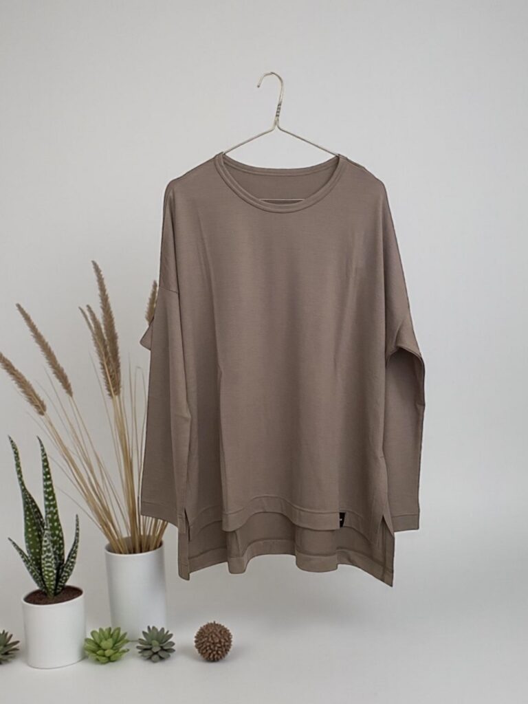 The Wool Jersey T-Shirt by Ujoh in grey, displayed on a minimal metallic hanger against a clean white background. The shirt's oversized silhouette, stepped hem, and soft wool jersey fabric are clearly visible, with green plants and wheat ears framing the left side of the composition.
