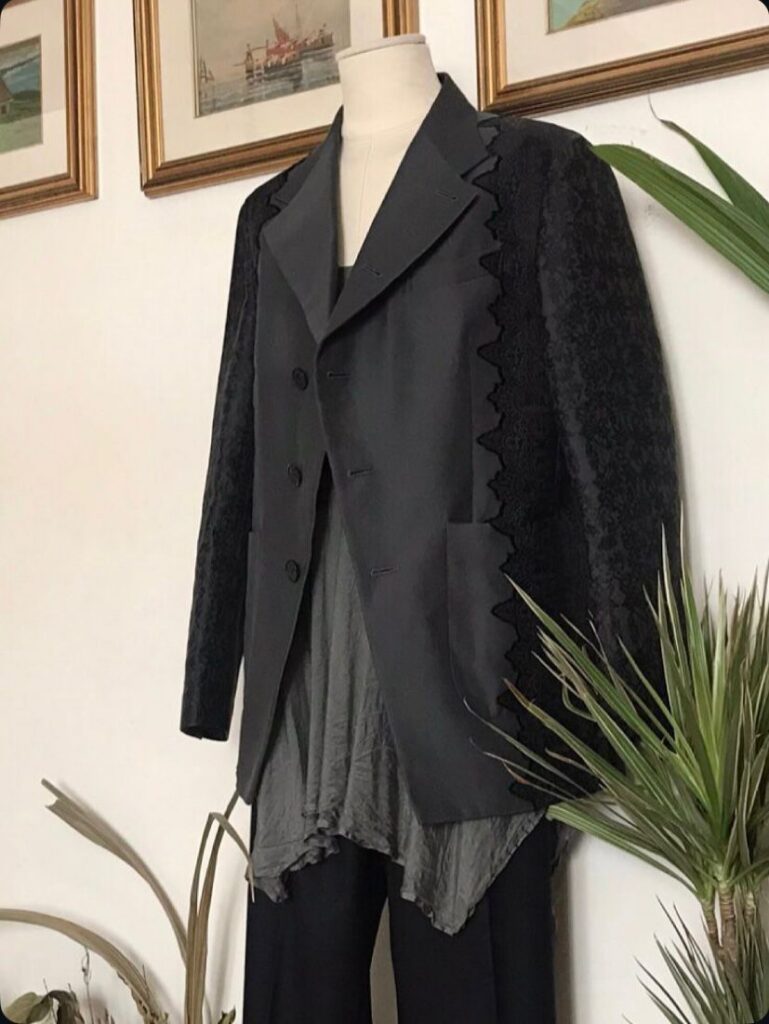 A tailoring mannequin wears The Lace Jacquard Blazer by Meagratia, layered over an asymmetric anthracite silk top and tailored wool pants. The scene is set against a clean white wall, framed by green plants on either side and a row of small paintings along the top, creating a minimalist and intentional studio atmosphere.