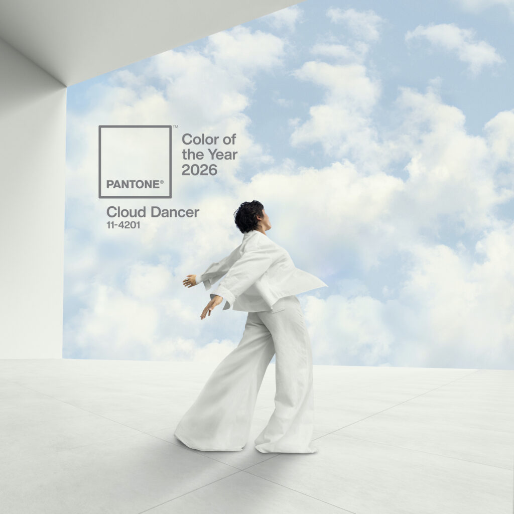 A conceptual image introducing Pantone's 2026 Color of the Year, Cloud Dancer. A woman in a flowing garments of a tranquil shade moves gracefully. She is posed as if dancing or floating effortlessly between wispy clouds and a smooth, reflective white floor. The overall scene evokes a feeling of lightness, calm, and ethereal movement.