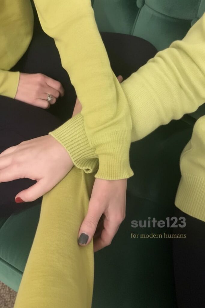 Celebrating twenty years of suite123. Three arms intertwined, wearing the same chartreuse knit, seated on a green velvet sofa. A portrait of connection between the two of us from suite123 and a close friend.