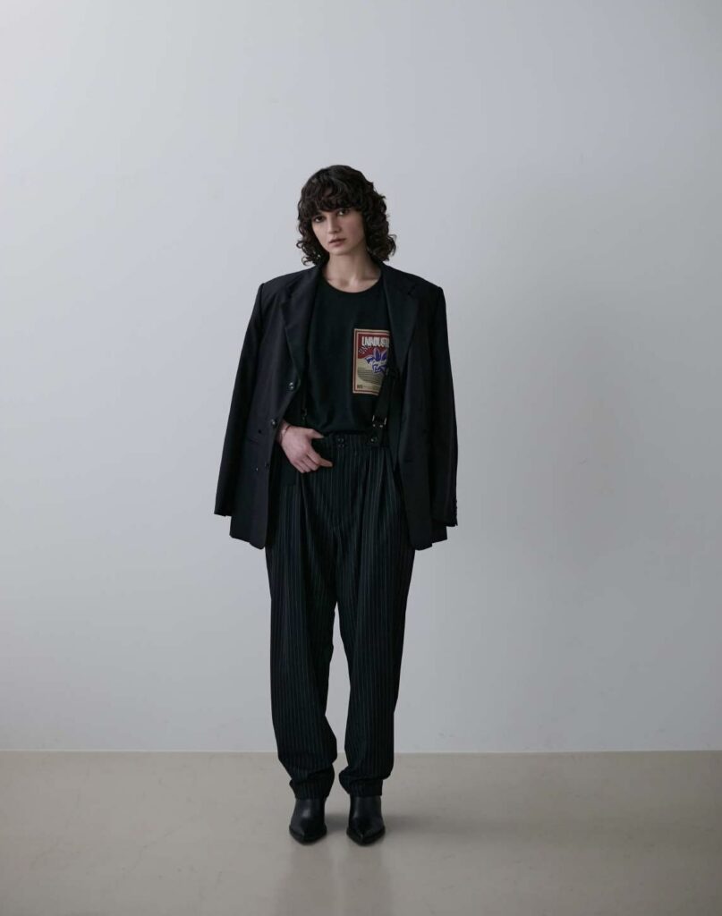 Curly-haired brunette models The Suspenders Trousers by Meagratia — black pinstripe trousers with integrated suspenders — paired with a black t-shirt, a black blazer draped over the shoulders, and black leather booties. Neutral grey studio background.