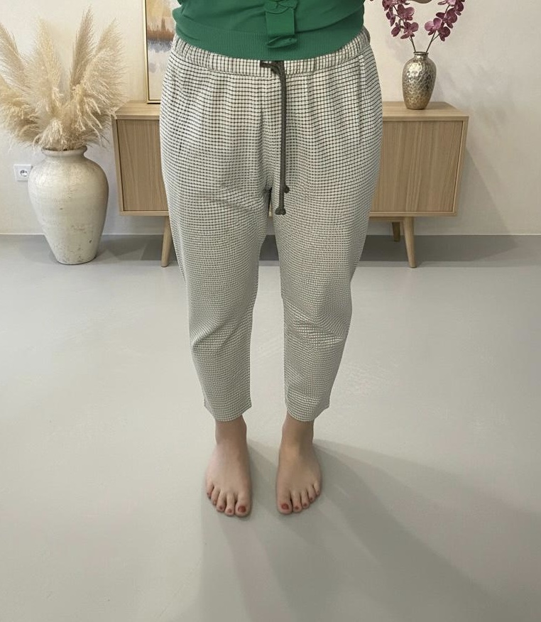 The Check Trousers by GoodNeighbors Shirts shown from waist to bare feet, revealing the distinctive burgundy-and-dark-green check on crisp white cotton. Set against a minimalist backdrop of warm wooden wall and table, adorned with two vases and understated decor.