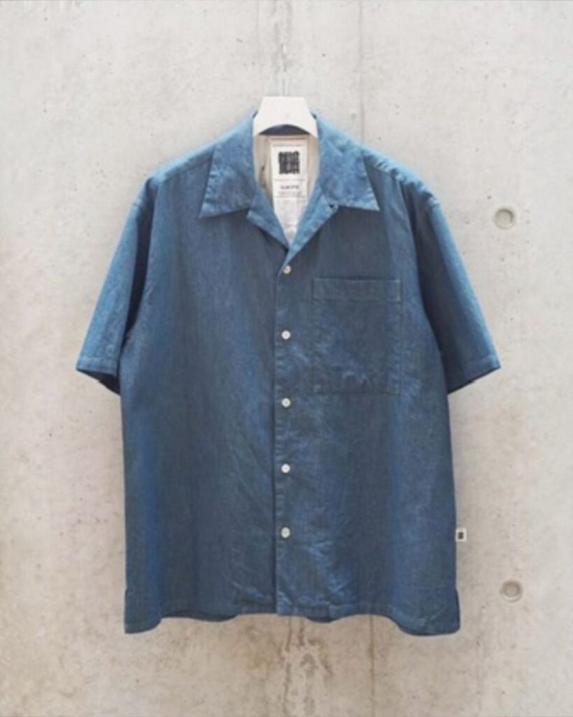 The Clay Indigo Short Sleeve — naturally dyed cotton shirt by GoodNeighbors Shirts. Suspended against a muted grey backdrop. Earth-born colour, tailored ease.