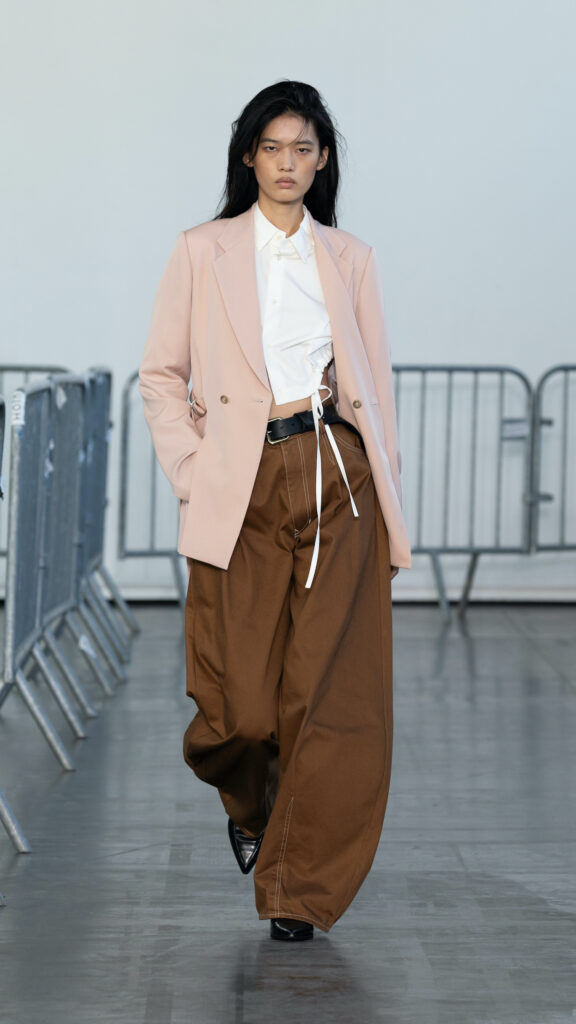 Model on the Ujoh SS27 Paris runway demonstrating the collection's tailoring meets sportswear concept through a powder pink blazer layered over a white shirt and balloon terracotta jeans.